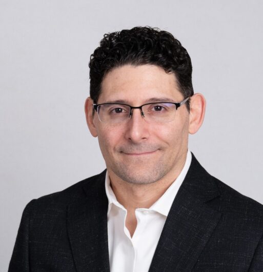 A man with short, curly dark hair and glasses wearing a black suit jacket and white shirt poses in front of a plain light background.