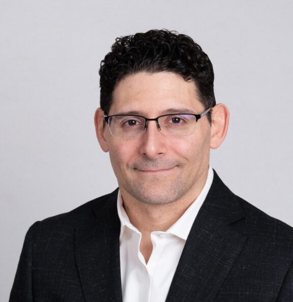 A man with short, curly dark hair and glasses wearing a black suit jacket and white shirt poses in front of a plain light background.