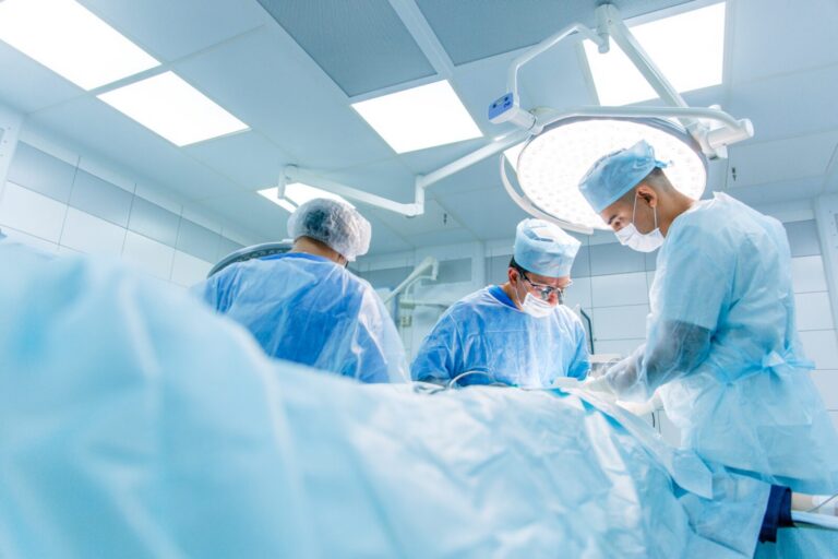 Three surgeons in scrubs and masks perform an operation under bright surgical lights in a sterile operating room.