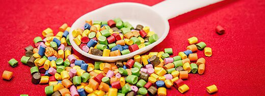 A white plastic spoon with colorful plastic pellets scattered on a red surface.
