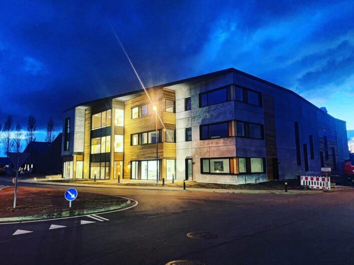 A modern three-story building with illuminated windows is pictured at dusk, surrounded by a road and minimal landscaping under a dark, cloudy sky.