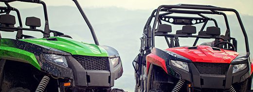 Two off-road utility vehicles, one green and one red, parked side by side with a blurred outdoor background.