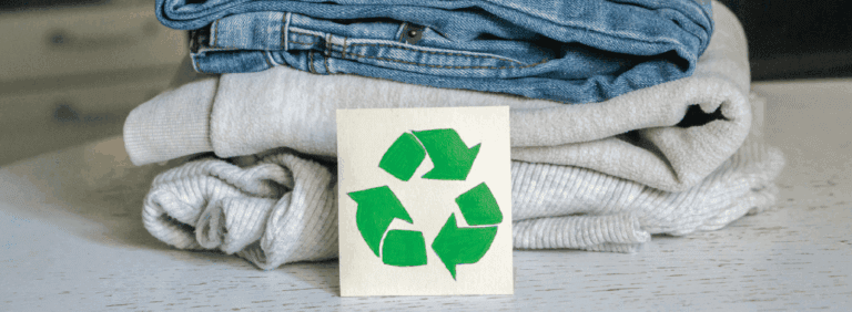 A stack of folded clothes with a paper showing a green recycling symbol placed on top, set on a light-colored surface.