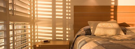Sunlight streams through window blinds onto a neatly made bed with pillows, next to a wooden nightstand in a cozy bedroom.