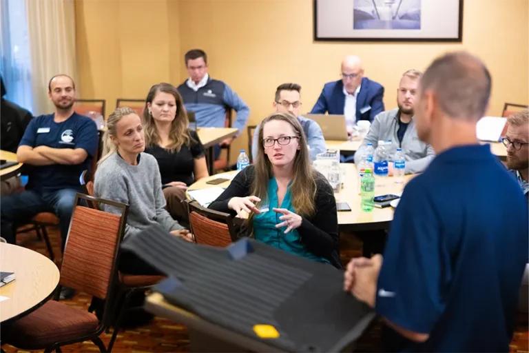 A group of people sit around tables in a conference room, listening to a speaker at a podium; one woman gestures while speaking.
