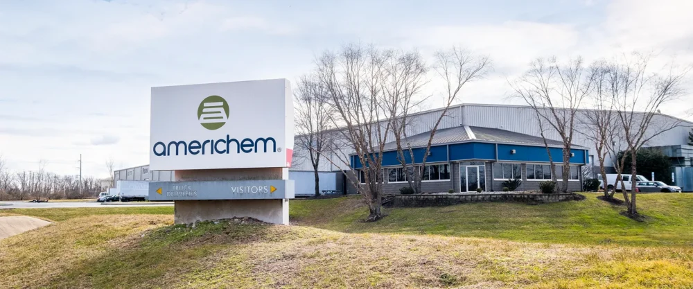 A large sign reading "Americhem" stands in front of a modern industrial building with blue and gray exterior panels, surrounded by leafless trees.