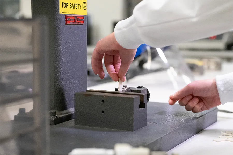 A person operates a machine in a lab, positioning a small sample in a metal clamp labeled "FOR SAFETY.