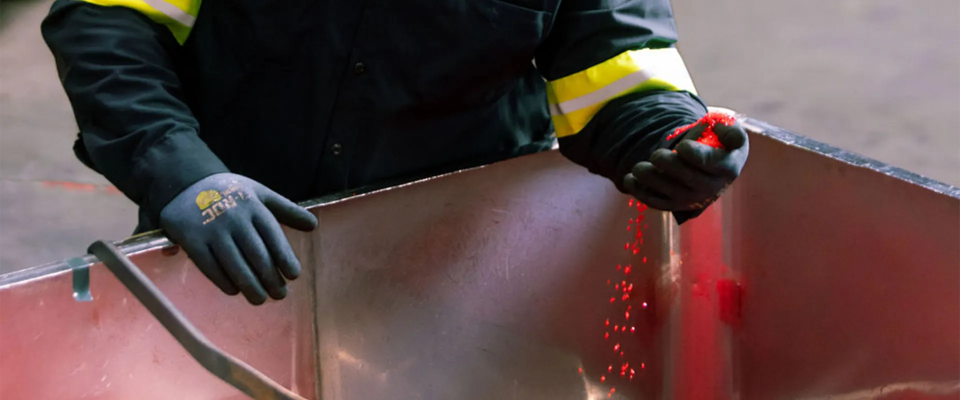 A worker in dark protective clothing and gloves pours red granules from one hand into a large metal container.