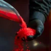 A person wearing black gloves holds red plastic pellets as more are poured from a metal scoop.