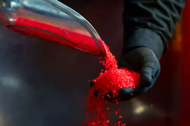 A gloved hand holds red plastic pellets being poured from a metal scoop.