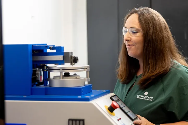 A woman in safety glasses operates a blue scientific instrument in a lab, wearing a green shirt with an "omanchem" logo.