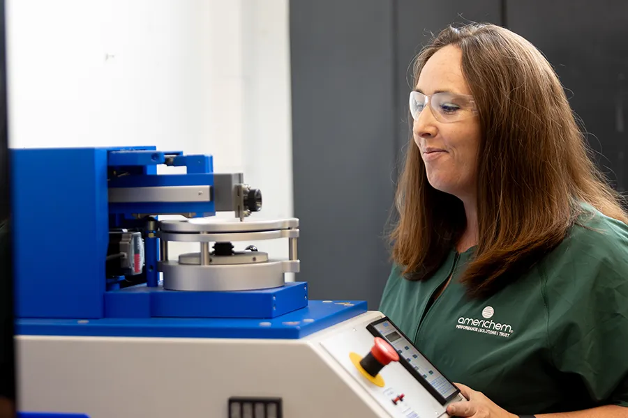 A woman in safety glasses operates a blue scientific instrument in a lab, wearing a green shirt with an "omanchem" logo.