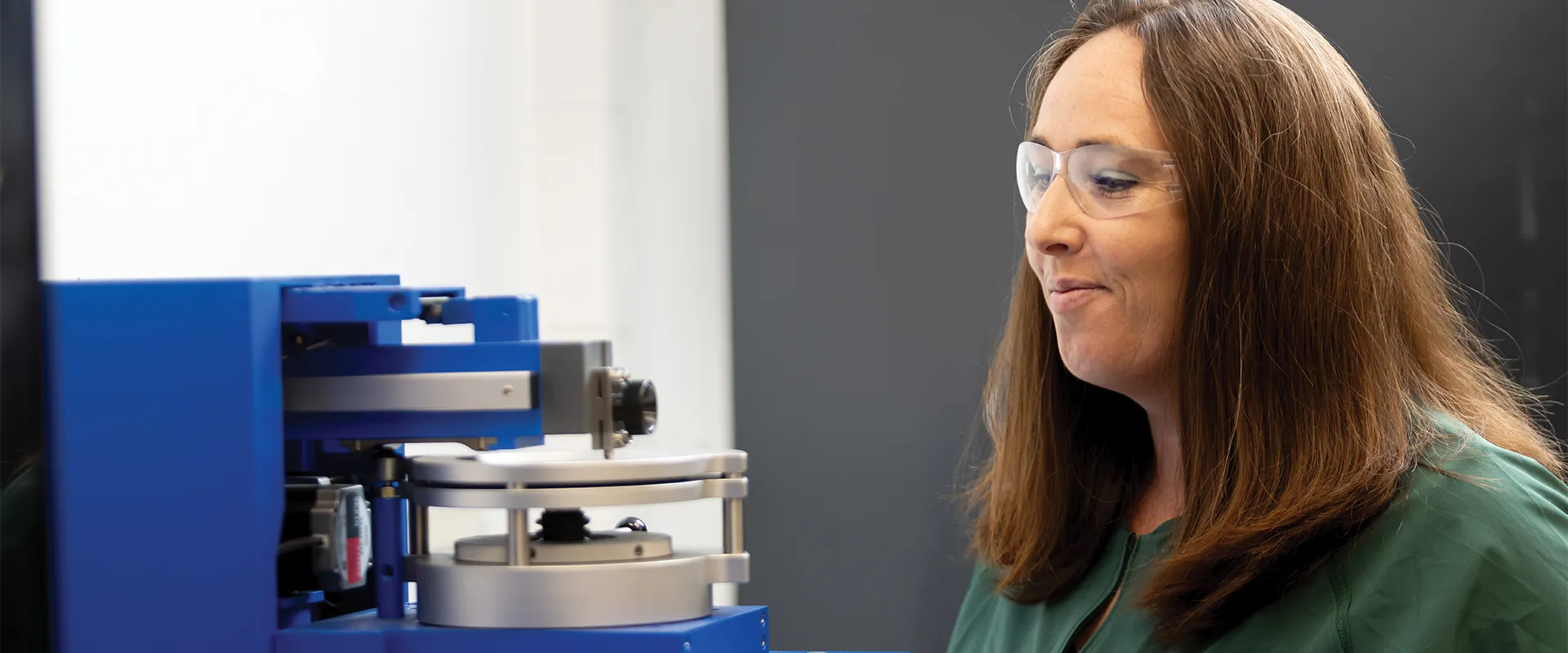 A woman wearing safety glasses operates a blue laboratory instrument in a lab setting.