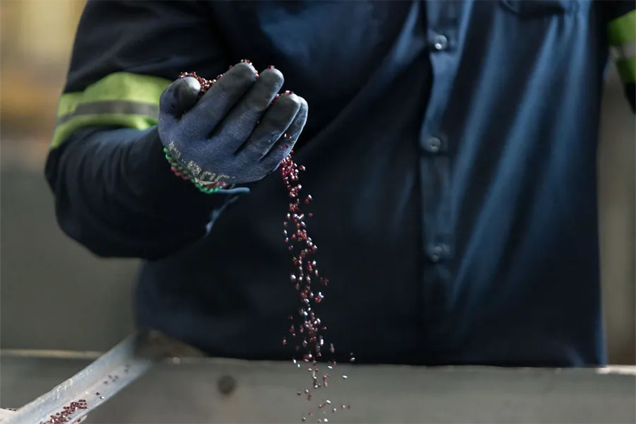 A person wearing a blue work shirt and gloves lets small red granules fall from their hand over a metal surface.