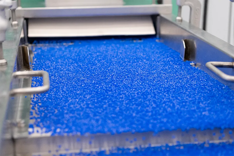 Blue plastic resin pellets move along an industrial conveyor belt in a manufacturing facility.