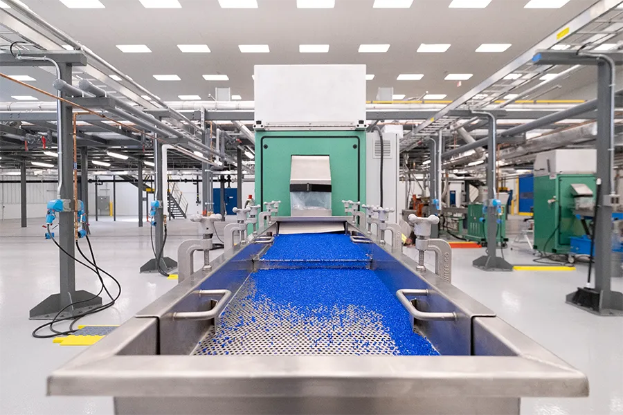 View of an industrial facility with a conveyor system transporting blue plastic pellets toward a green processing machine in a clean, brightly lit environment.
