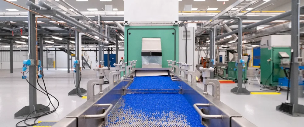 Blue plastic pellets move along a conveyor belt in a brightly lit industrial facility with various machinery and pipes in the background.