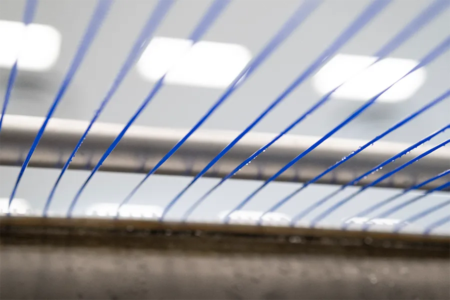 Close-up of several parallel blue threads running across a metallic frame in a brightly lit industrial or manufacturing setting.