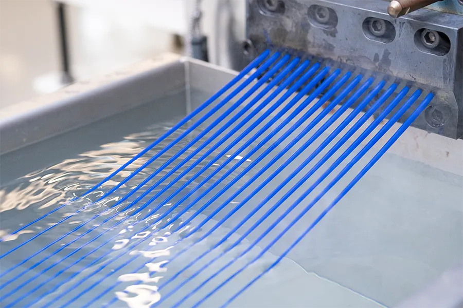 Bright blue synthetic fibers are extruded from a metal machine and extend into a water bath, likely part of an industrial manufacturing process.