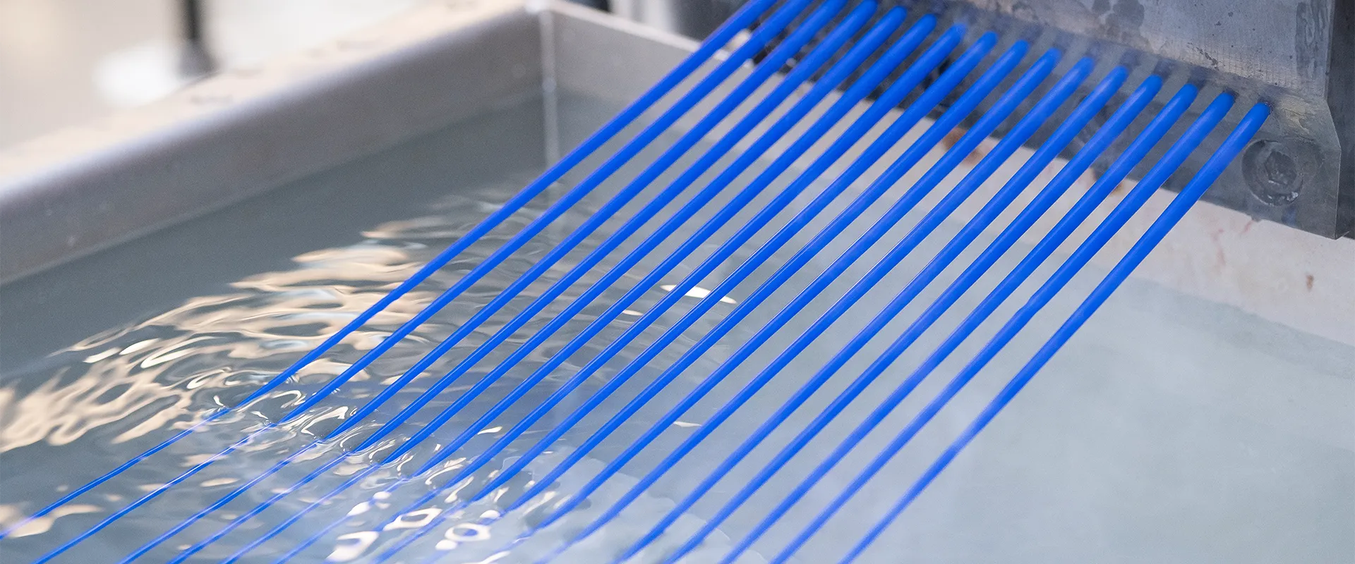 Several blue synthetic fibers are being extruded from a machine into a water bath, likely as part of a manufacturing or cooling process.