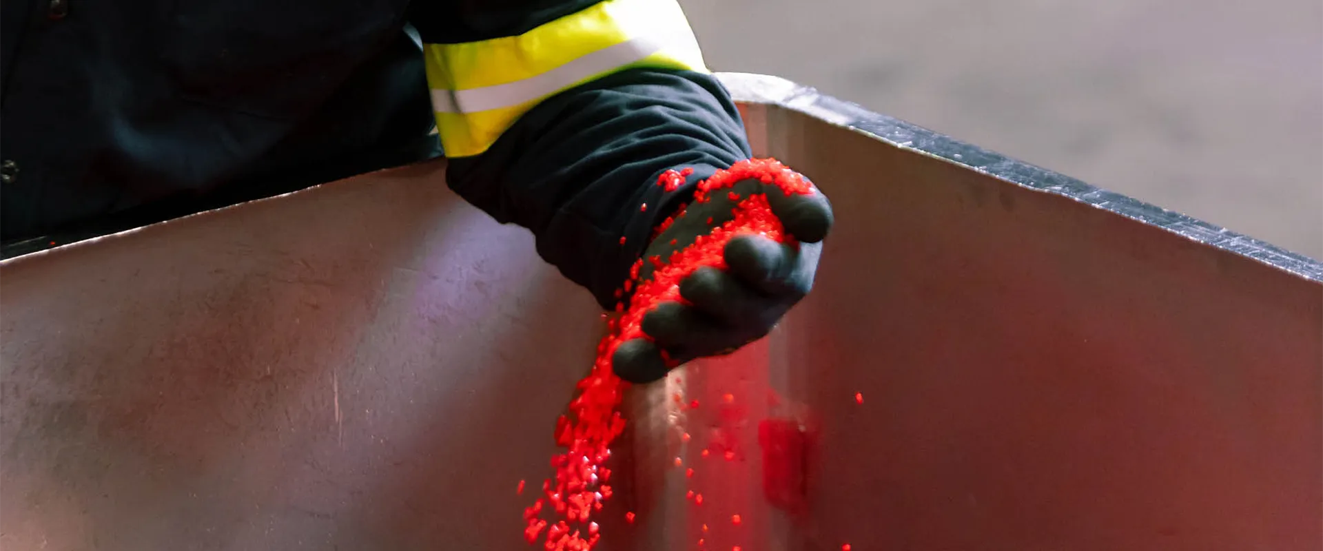 A person wearing a black glove pours red plastic pellets from their hand into a large metal container.
