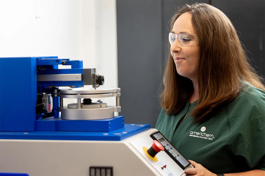 A woman in safety glasses operates a blue laboratory testing machine, wearing a green Americhem-branded shirt and holding a touchscreen control panel.