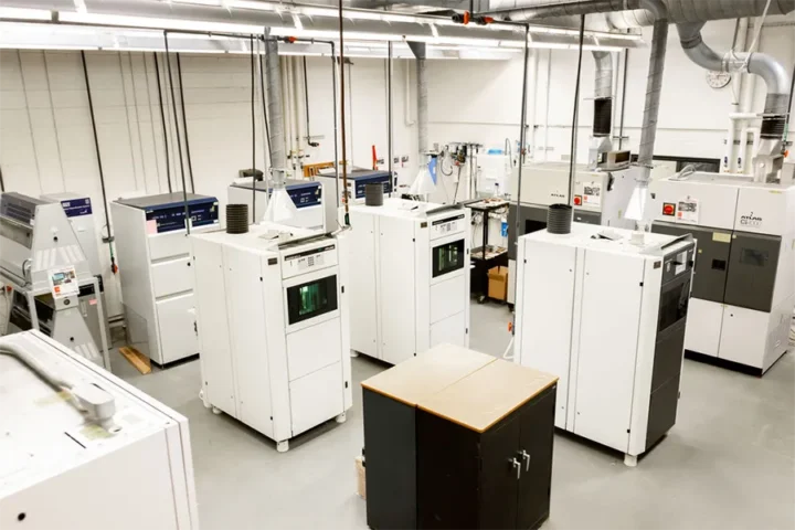 A clean, organized lab with multiple large industrial 3D printers and equipment arranged in rows, white walls, and overhead fluorescent lighting.