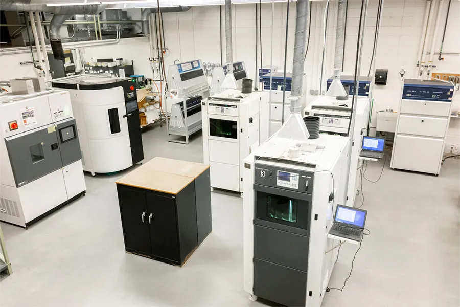 A laboratory room filled with several large 3D printers and related equipment, with a black cabinet in the center and computers connected to some machines.