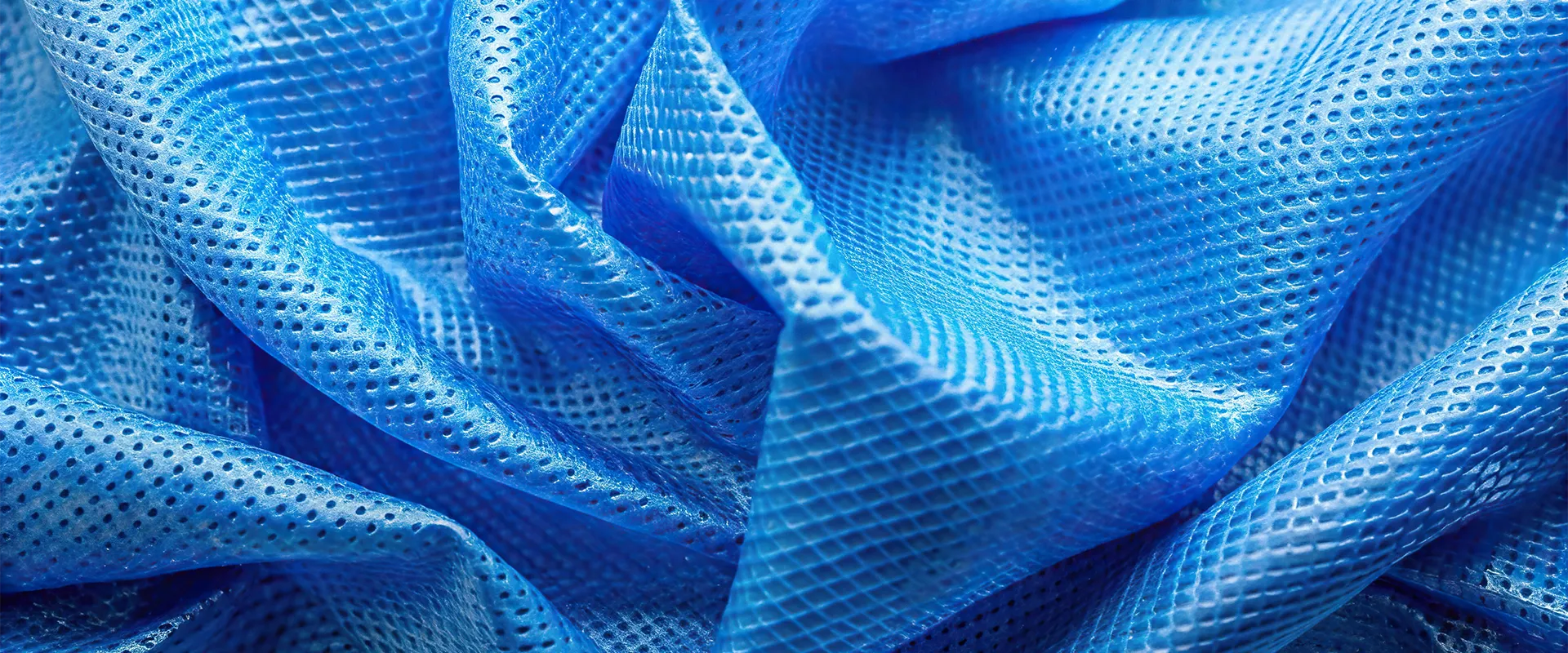 Close-up of textured, blue, perforated fabric with a grid and mesh pattern, showing multiple folds and light reflecting off the surface.