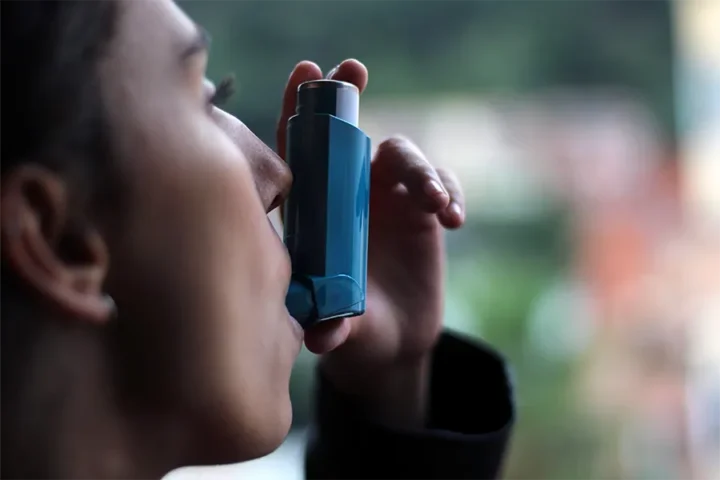 A person uses a blue inhaler, holding it to their mouth, with a blurred outdoor background.