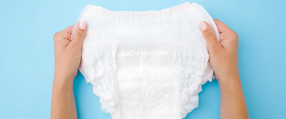 Two hands holding a white disposable adult diaper against a blue background.