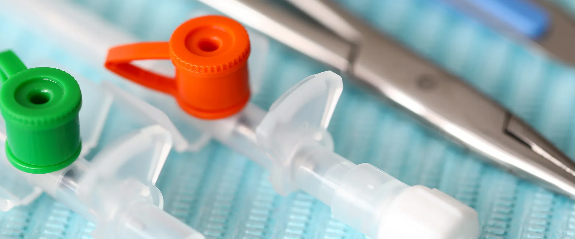 Close-up of medical cannulas with green and orange caps on a blue surface, with surgical scissors in the background.