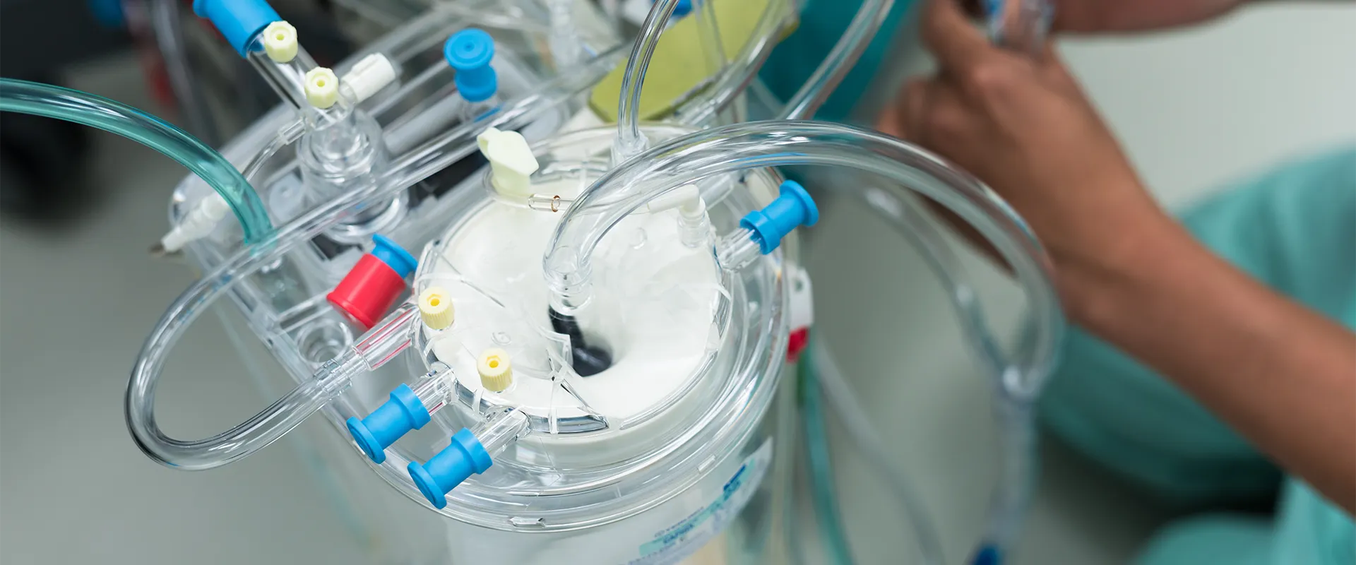 A close-up of a medical device with multiple tubes and connectors, likely part of a heart-lung machine, with a person's hands adjusting equipment in the background.