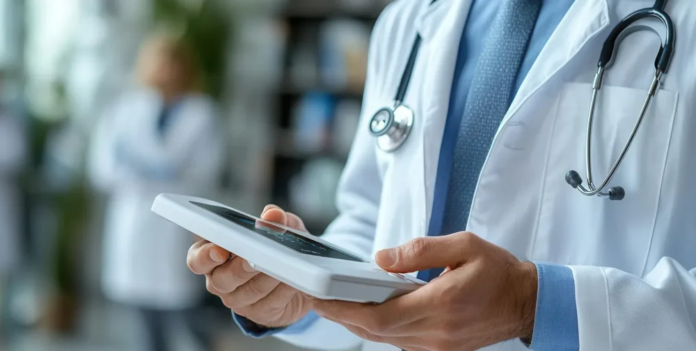 A doctor in a white coat with a stethoscope around the neck holds a tablet device; a blurred medical professional is visible in the background.