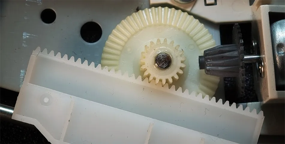 Close-up of interlocking plastic gears and a white rack inside a mechanical device, showing gear teeth and assembly components.