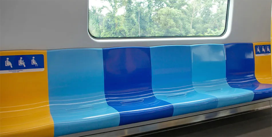 Empty seats in a train carriage with alternating blue and yellow colors, next to a large window with trees visible outside.