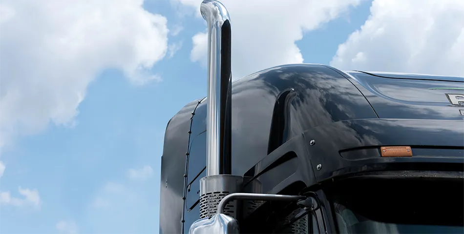Close-up of a shiny chrome exhaust pipe on the side of a black semi-truck cab against a cloudy blue sky.