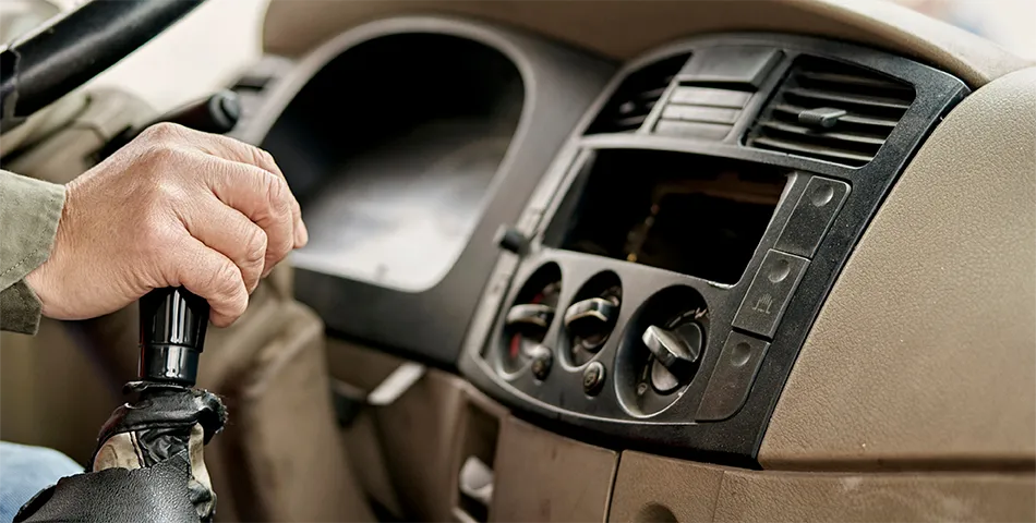 A person’s hand holding a manual gear shift lever inside a car, with the dashboard, air vents, and control knobs visible.