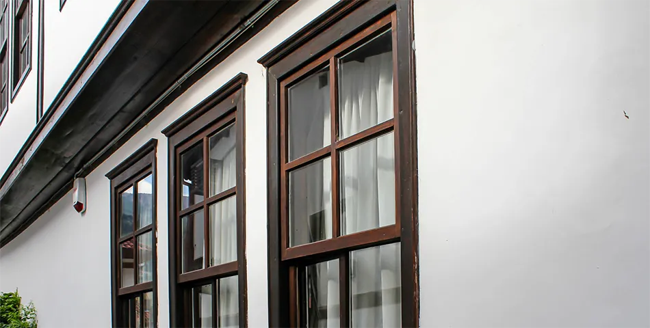 Three large rectangular windows with dark wooden frames are set into a white exterior wall, each window covered with white curtains inside.