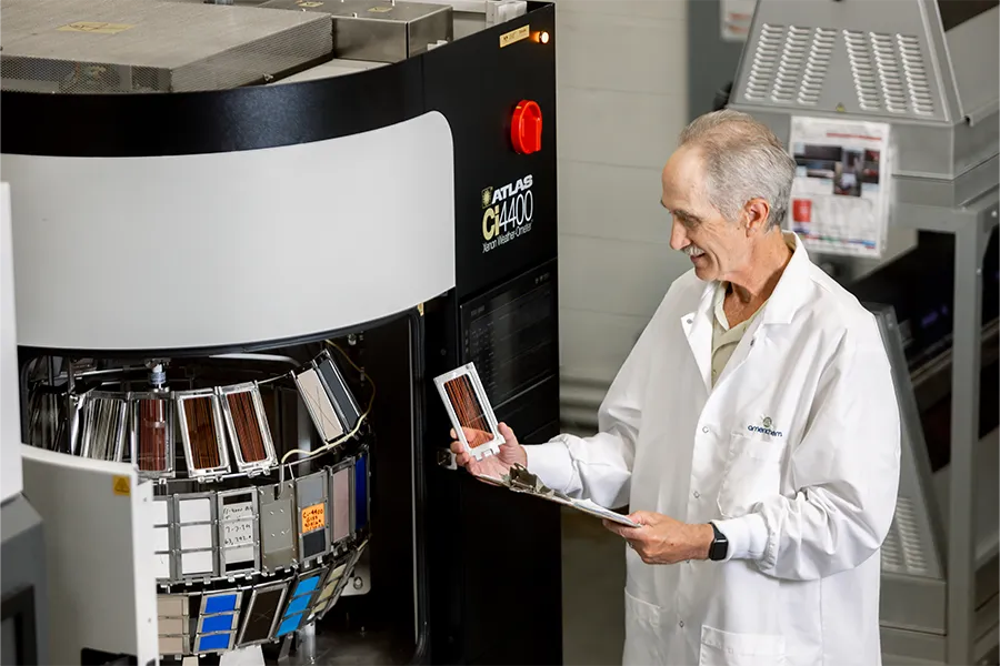 A scientist in a lab coat examines solar cell samples in front of a large testing machine, holding a clipboard and one of the samples.