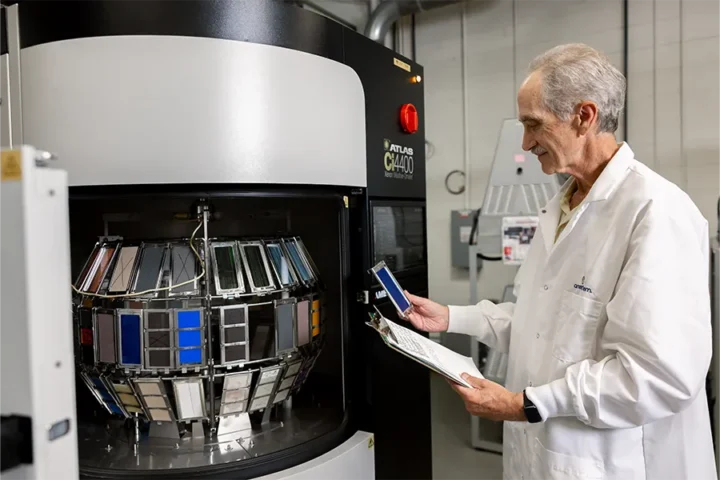 A technician in a lab coat inspects color sample panels inside a large testing machine, holding a clipboard and a sample for comparison.