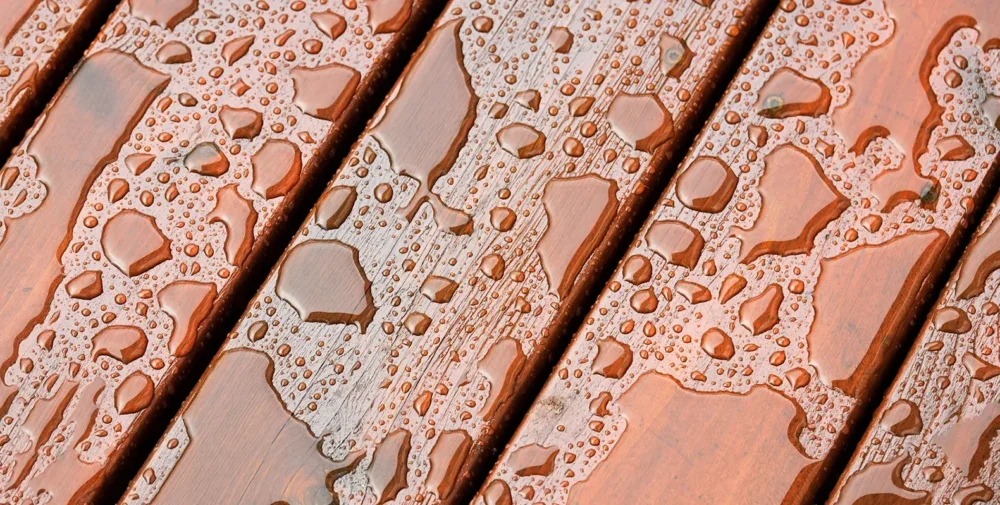 Close-up of water droplets and puddles on the surface of wooden planks, showing the texture and grain of the wood.