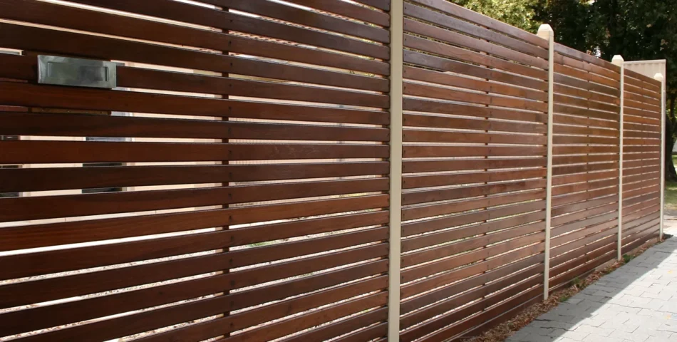 Horizontal wooden slat fence with evenly spaced boards and cream-colored vertical posts, bordering a paved walkway with greenery visible in the background.