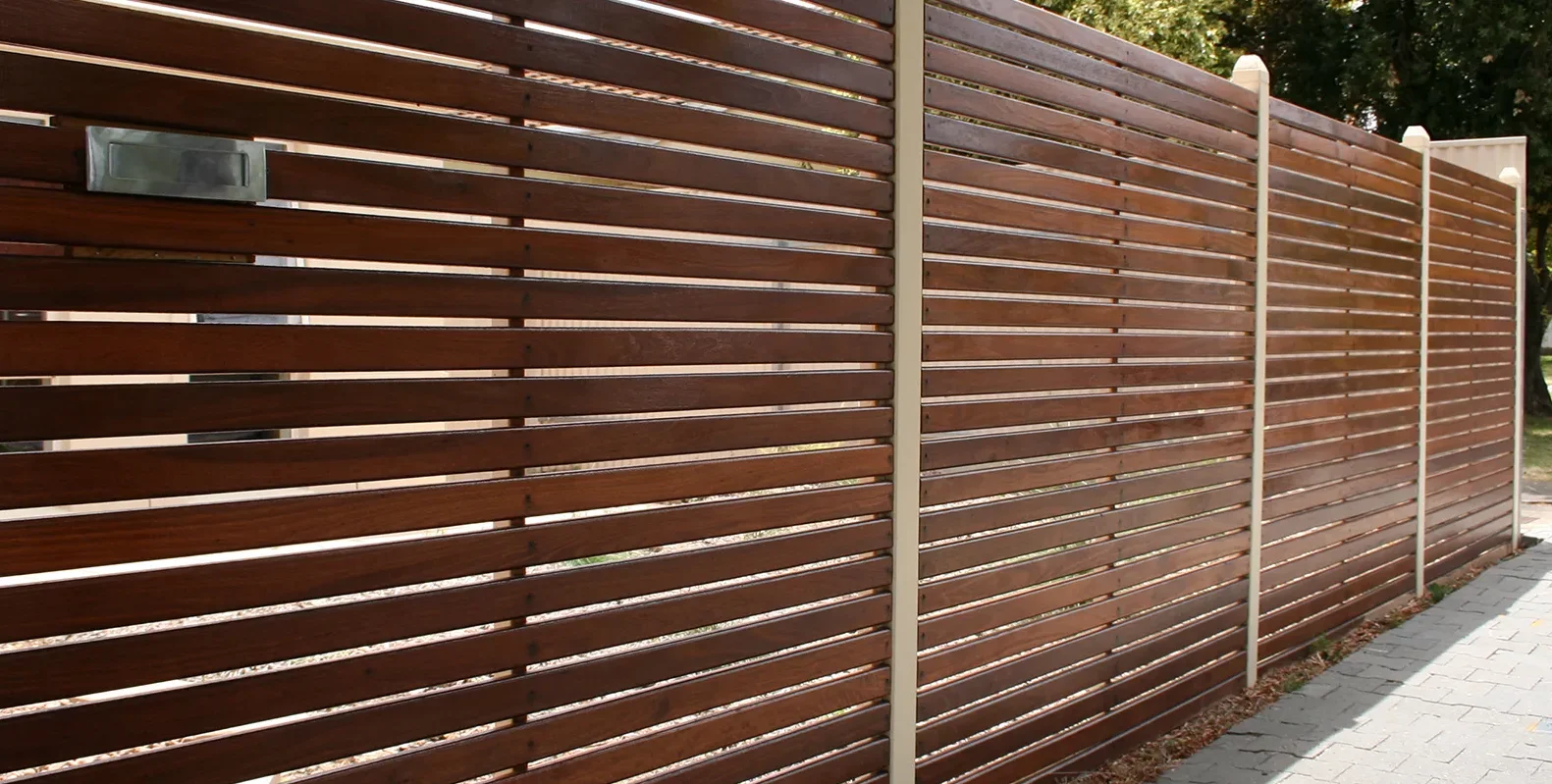 Horizontal wooden slat fence with evenly spaced boards and cream-colored vertical posts, bordering a paved walkway with greenery visible in the background.