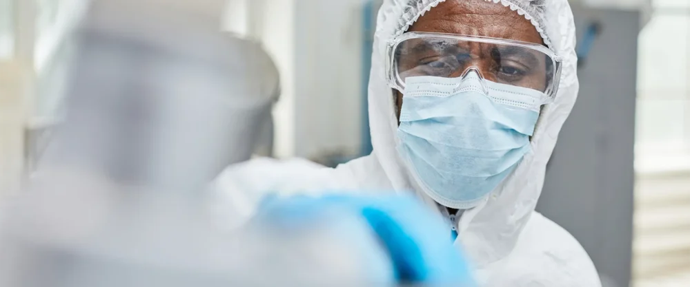A scientist wearing protective clothing, face mask, and safety goggles works with laboratory equipment.