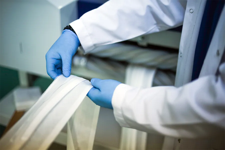 Person wearing blue gloves and a white lab coat handling strips of white fabric near industrial machinery.