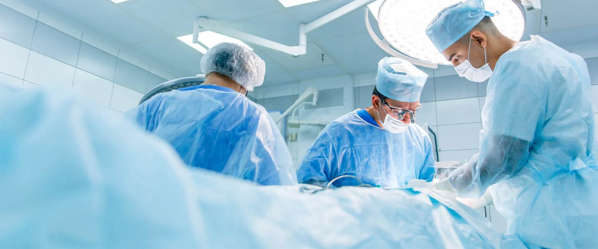 Three surgeons in scrubs and masks perform surgery in a brightly lit operating room with medical equipment visible in the background.