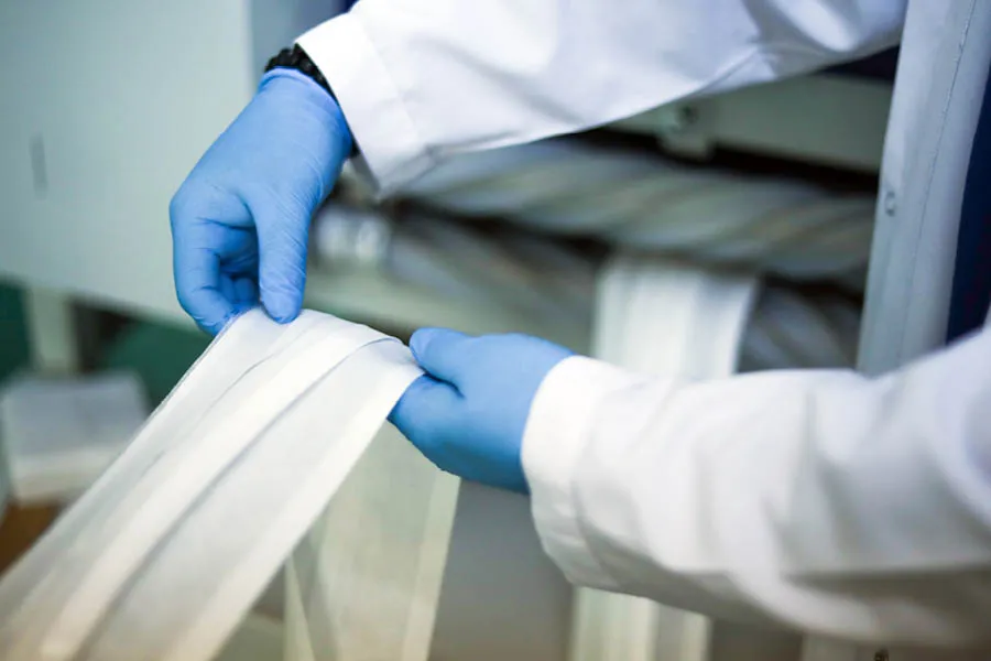 Person wearing blue gloves and a white lab coat handling strips of white fabric or material in an industrial or laboratory setting.