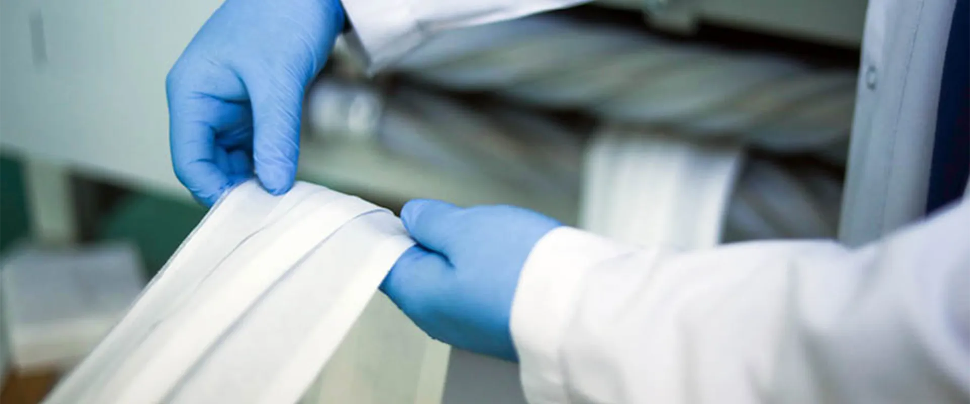 A person wearing blue gloves and a white lab coat inspects or handles strips of white fabric or material in an industrial or laboratory setting.