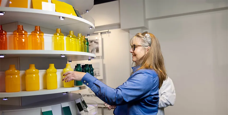 A woman in a blue shirt arranges colorful bottles on a display shelf, organized by color from yellow to green.